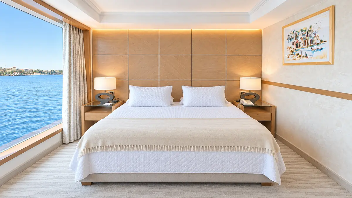 Amwaj Boat Renovation – Cabin with padded headboard wall, white bedding and a full-width window onto the Nile