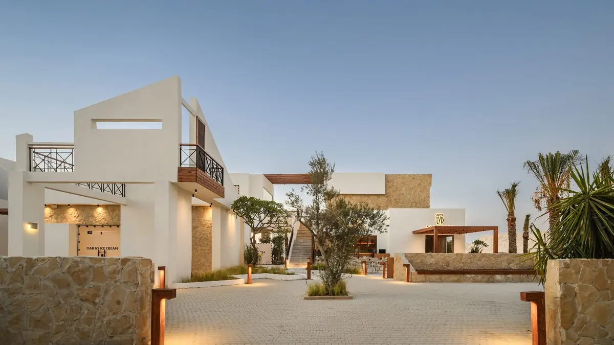 Masyaf Ras Al-Hekma – Community hub plaza at dusk with stone walls, olive tree and bollard lights