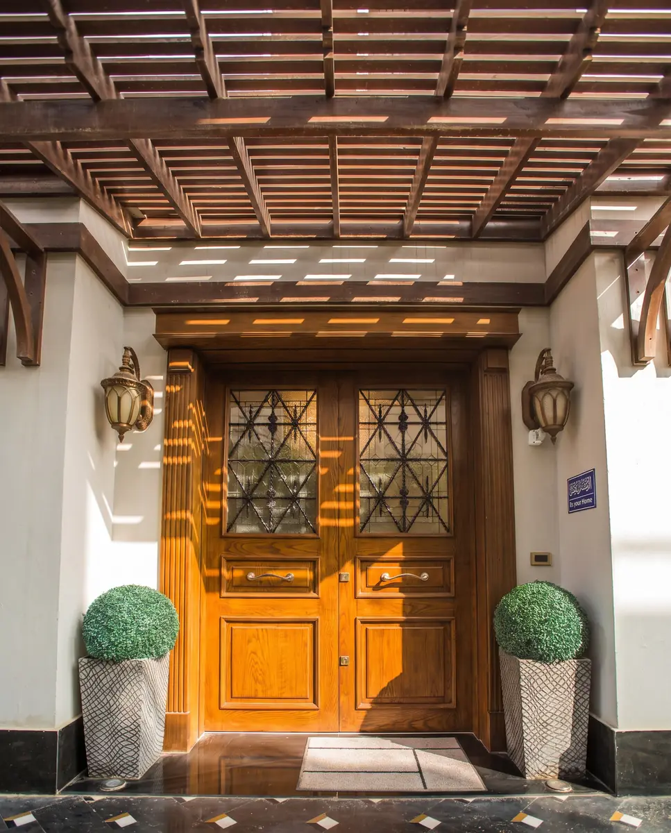 Mrs. M.A.'s Villa – Carved timber double door under a slatted pergola, flanked by lanterns and topiary