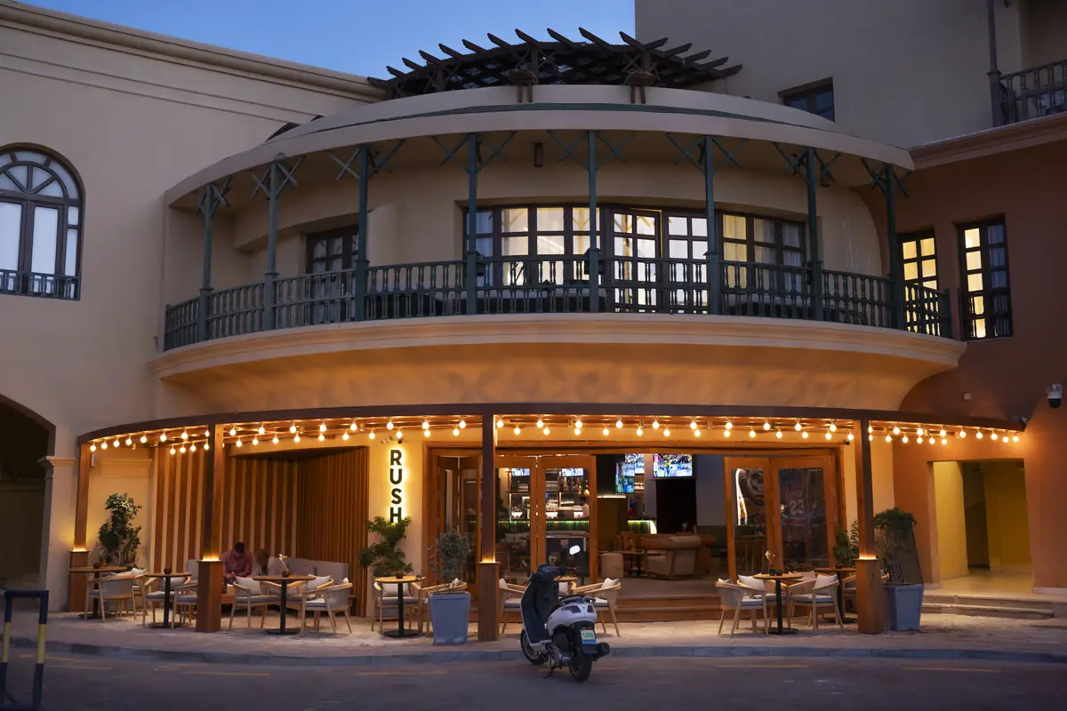 Rush – Sports Bar – Curved building façade at dusk with festoon-lit terrace below a wrought-iron balcony