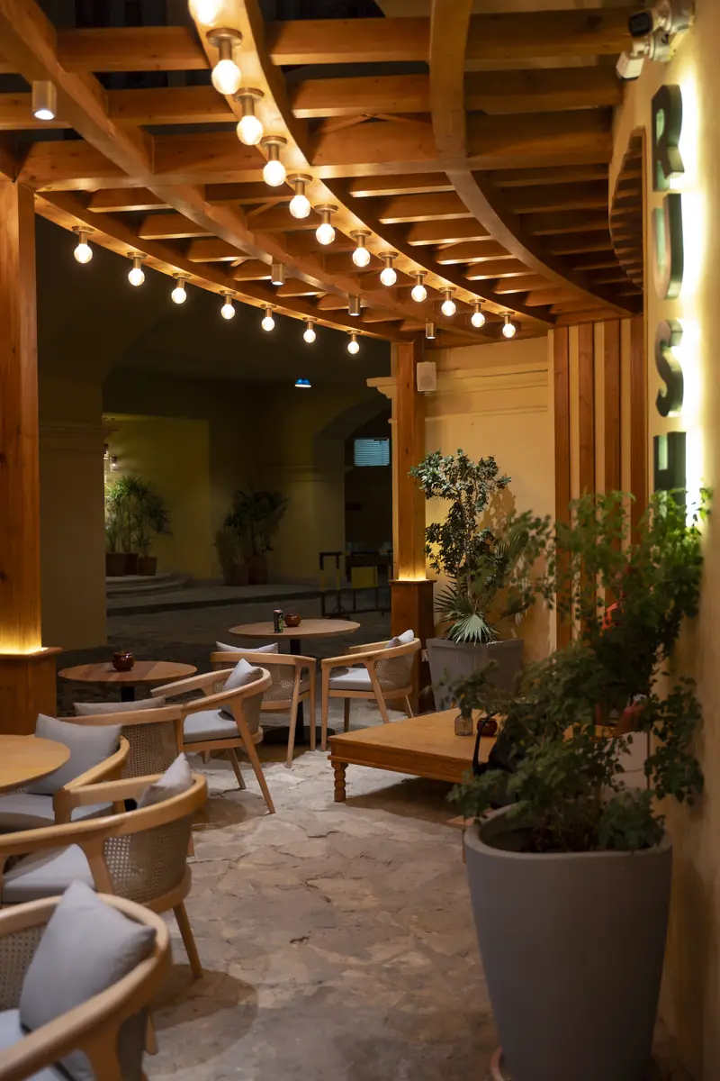 Rush – Sports Bar – Sitzbereich unter der Pergola mit Glühbirnenketten an Holzbalken und unregelmäßigem Natursteinboden