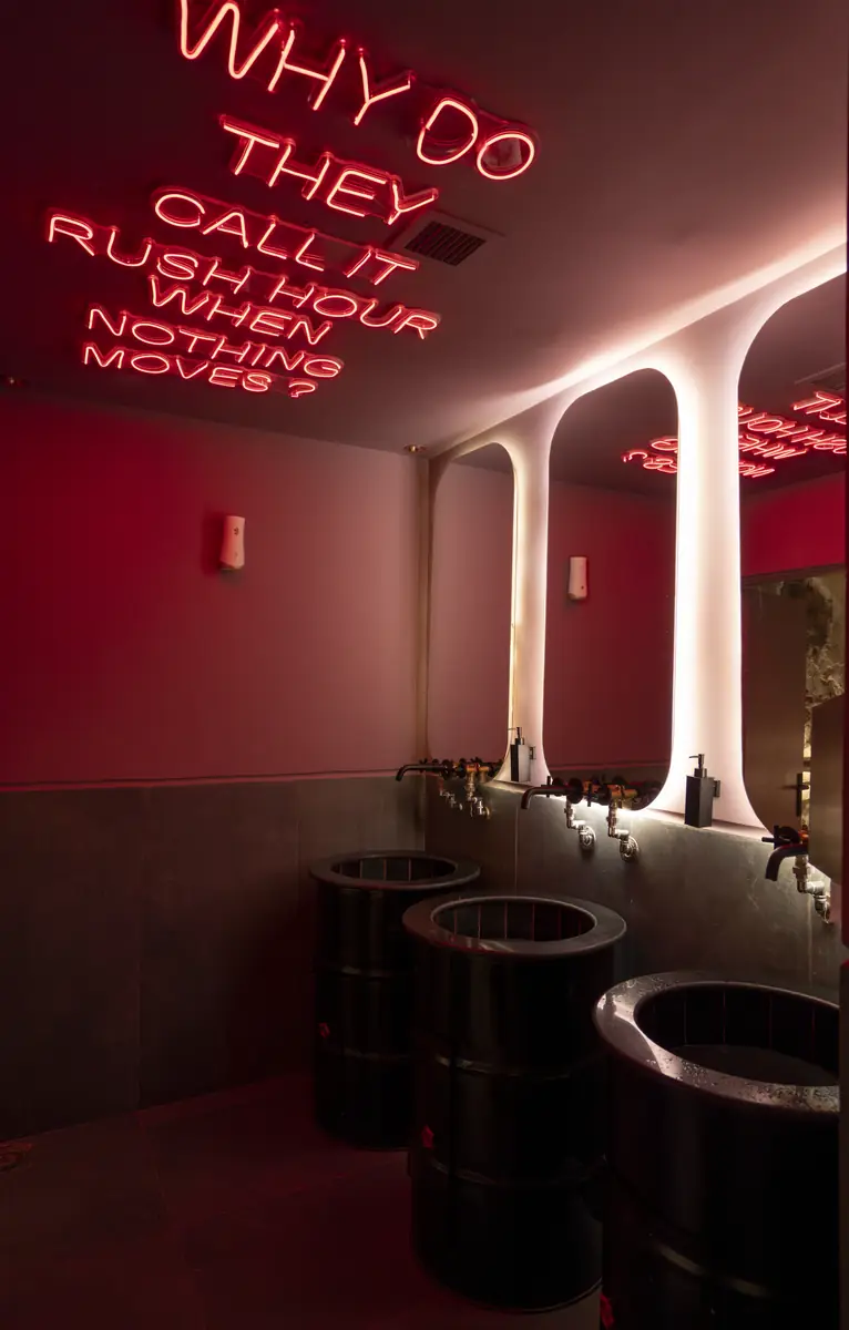 Rush – Sports Bar – Rot beleuchteter Waschraum mit Neon-Spruch an der Decke, Stahlfass-Waschbecken und hinterleuchteten Bogenspiegeln