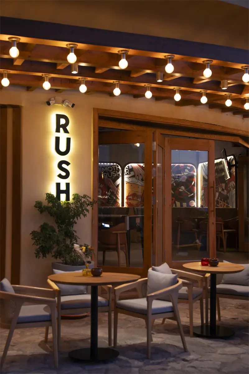 Rush – Sports Bar – Verglaster Eingang mit hinterleuchtetem senkrechtem Schriftzug, Bistrotischen und Lichterketten